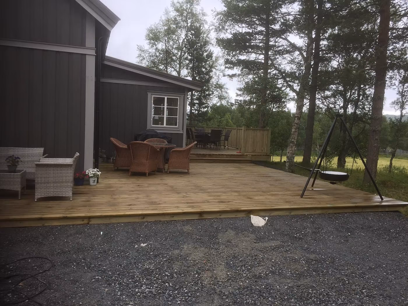 Ny terrasse i impregnert tre ved mørkbeiset hus, med hagemøbler og husketativ.