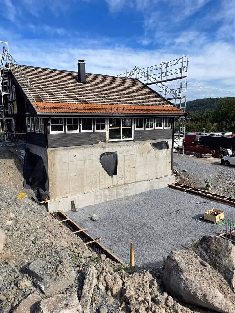 Byggeplass med oppføring av bolig i Bærum, fokus på takrenne, grunnarbeid og fundamentering, klar for videre bygging, omgitt av stein og grus, med byggeutstyr og stillas.