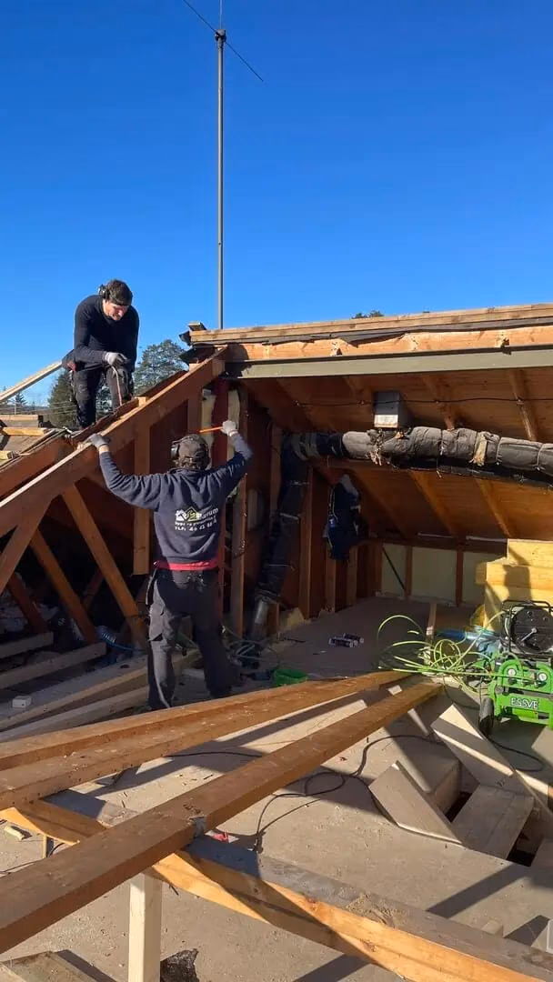 Bilde av to håndverkere som jobber med taktekking og utendørs konstruksjon, med byggematerialer og verktøy rundt dem, under en klar blå himmel.