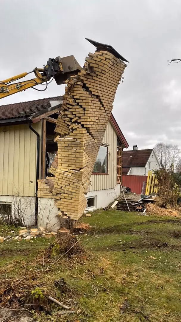 Bilde av en gravemaskin som river en gammel murvegg fra en bolig i Bærum, spesialisert i bygg- og graveservice, inkludert mur- og fundamentarbeid.