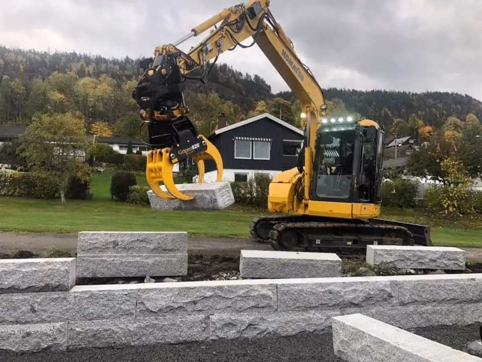 Gravemaskin som flytter store steinblokker for oppbygging av terreng og fundament, med fokus på profesjonell bygg- og graveservice i Bærum.