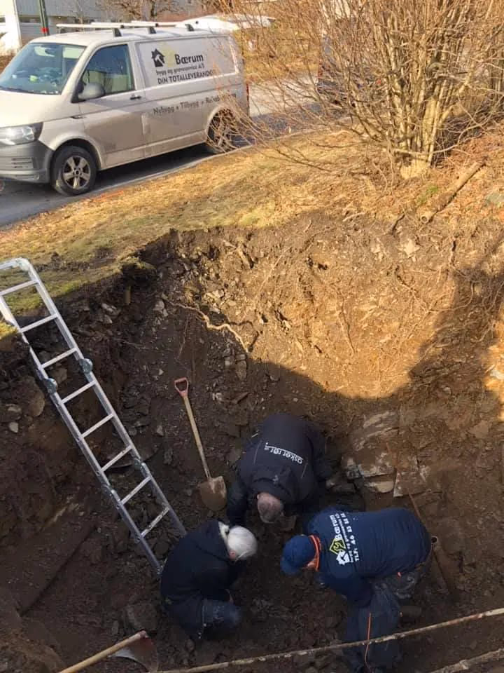 Bilde av gravearbeid utført av Bærum Bygg og Graveservice, med ansatte som jobber i en utgravningsgrøft, utstyr og verktøy for graving, med en firmebil i bakgrunnen.