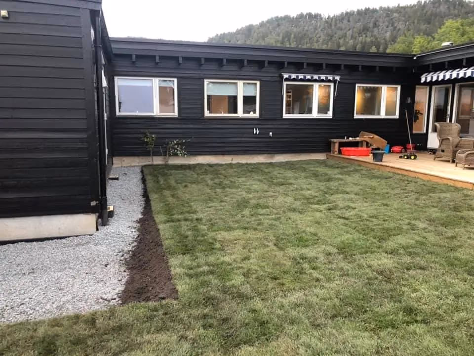 Velstelt hage med nyplenert gressplen, terrasse med utemøbler, og moderne svart husfasade, omgitt av naturskjønn skog i bakgrunnen.