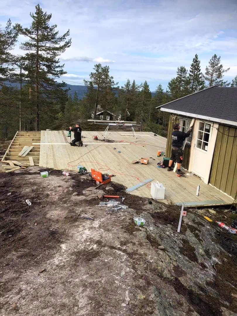 Bildet viser bygging av terrasse med treverk på et utendørs område i Bærum, med arbeidere som legger gulvplanker, omgitt av skog og fjell i bakgrunnen.