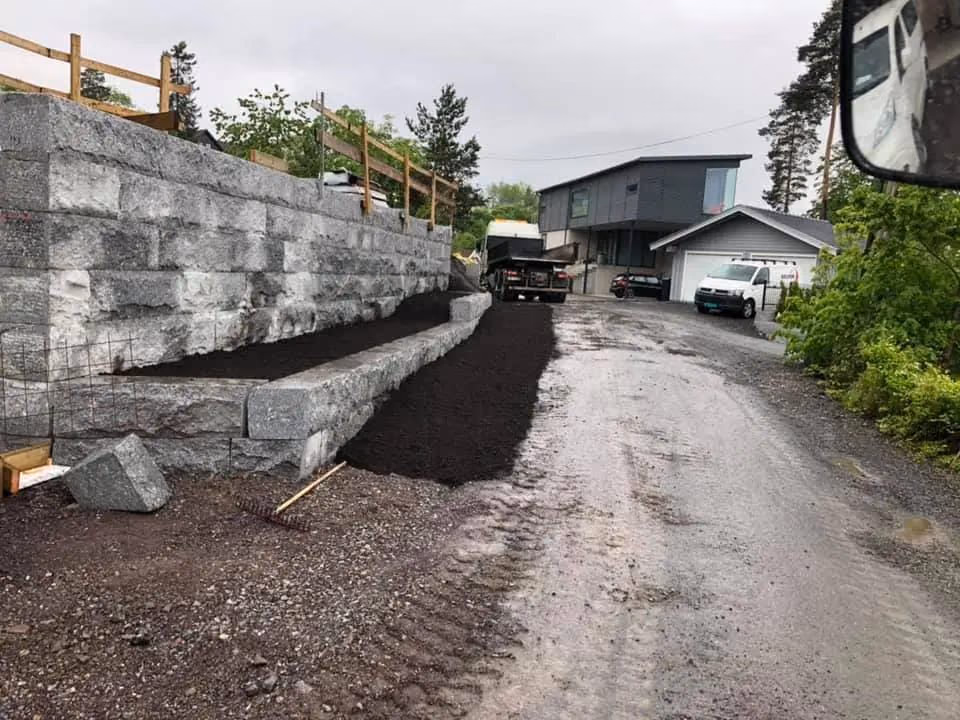 Bildet viser en opparbeidet tomt med støttemur av betongblokker, ny jord og grusvei, utført av Bærum Bygg og Graveservice, spesialisert på graving, tomteutforming og støttemurer.